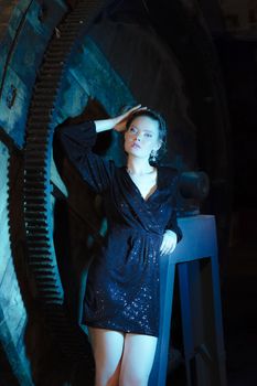 Fashion portrait of a woman in a sparkling black dress leaning against industrial machinery.