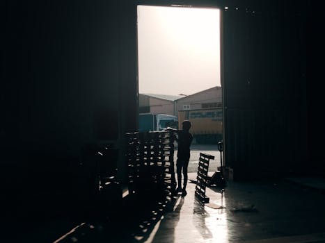 A worker arranges wooden pallets in a warehouse, backlit by sunlight streaming through an open door.