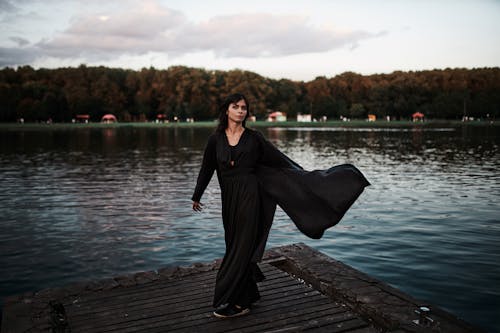 Woman in Black Dress Standing on Brown Wooden Dock near Body of Water