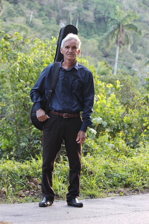 Free Portrait of an elderly musician standing outdoors with a guitar case, surrounded by greenery. Stock Photo