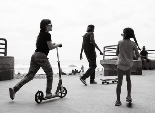 Free Children and a man enjoying leisure activities on a beachside promenade with scooters and skateboards in grayscale. Stock Photo