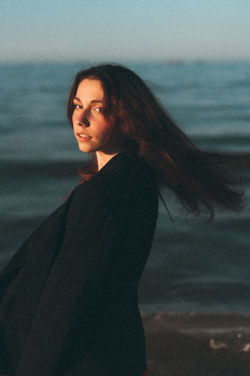 Free Stylish woman in a black coat gazes confidently at the camera with a serene sea background. Stock Photo