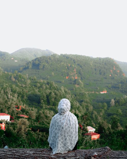 Free A woman in a patterned hijab sits on a log gazing at the lush countryside and mountains. Stock Photo