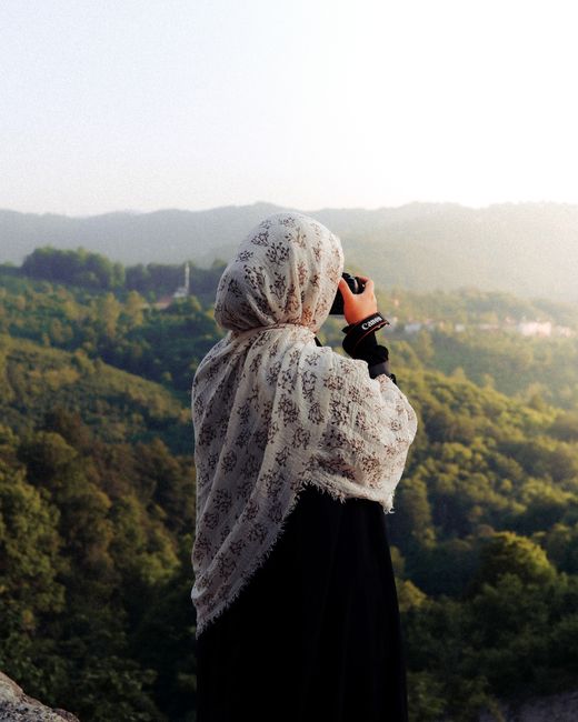 A woman in a hijab uses a camera to capture a beautiful mountain landscape.