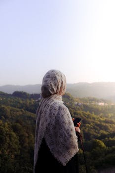 A woman in a hijab holds a phone while gazing at a lush green forest under a clear sky.