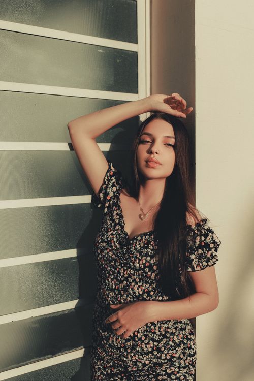 Free Elegant young woman in floral dress posing by a glass wall with serene expression. Stock Photo