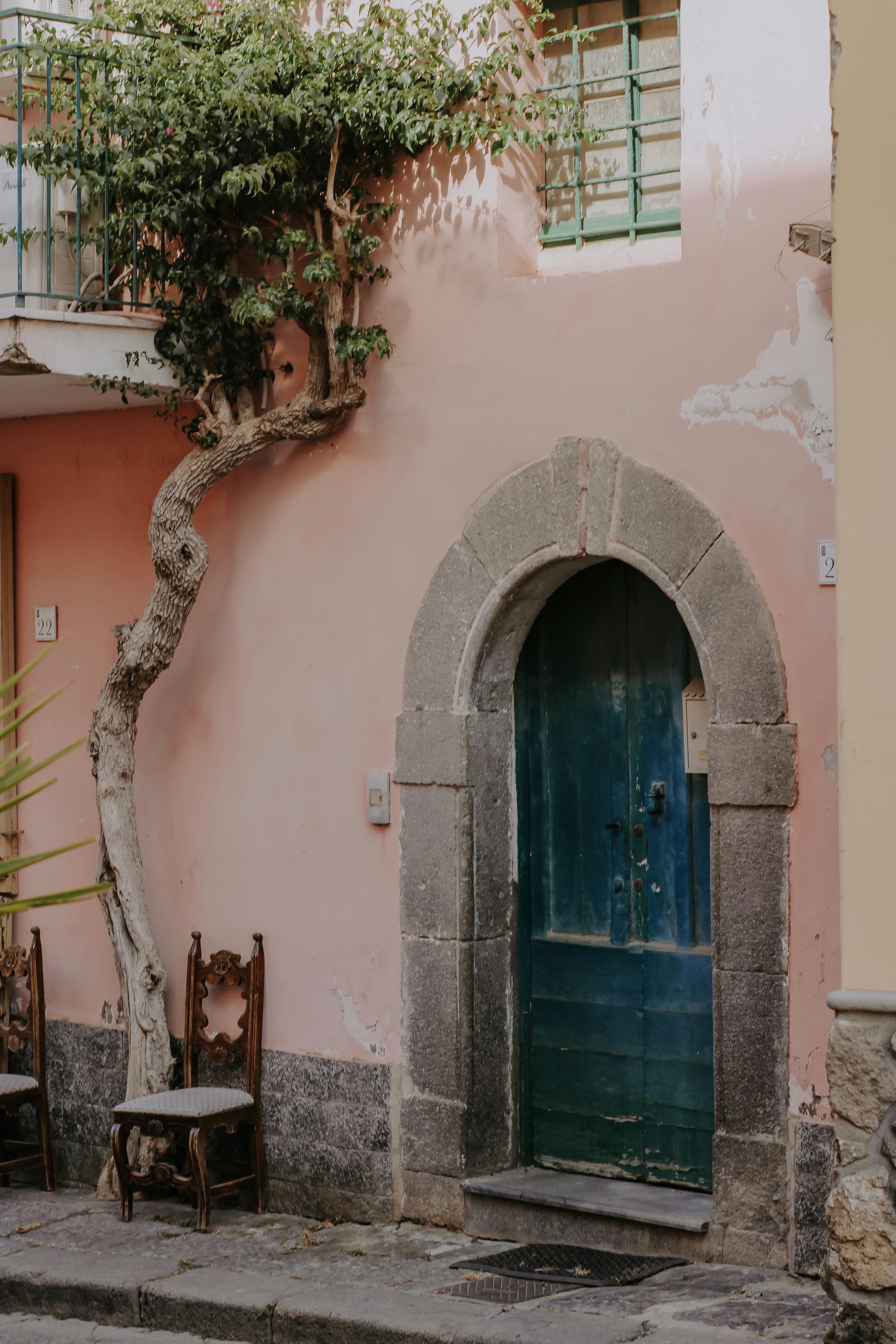 Free An elegant rustic door set in a pink plaster wall with ivy and wooden chairs. Stock Photo
