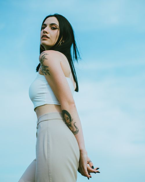 Free Confident woman with tattoos wearing white top posing thoughtfully against a clear blue sky. Stock Photo