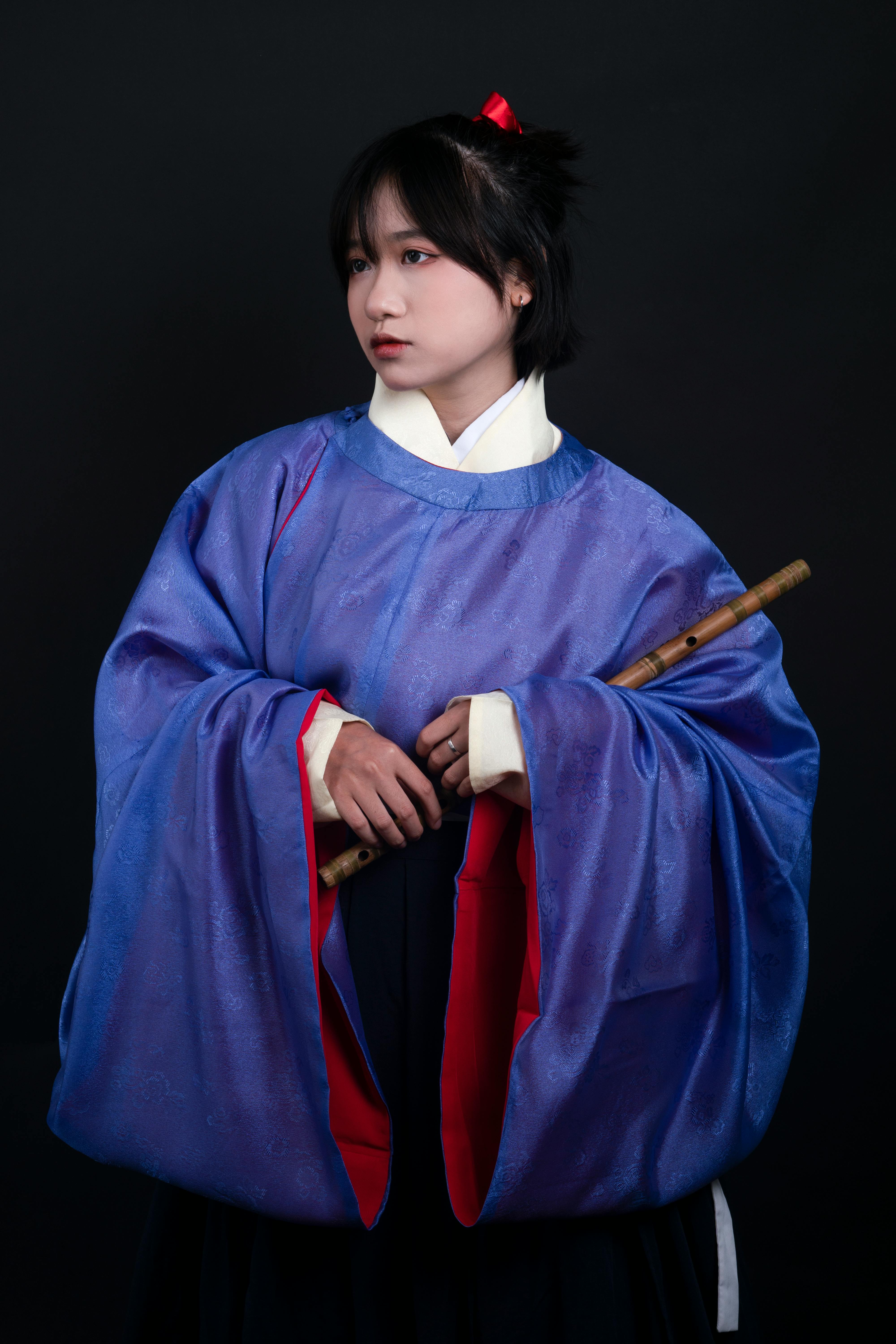 Portrait of an East Asian woman in traditional blue dress holding a flute against a dark backdrop.