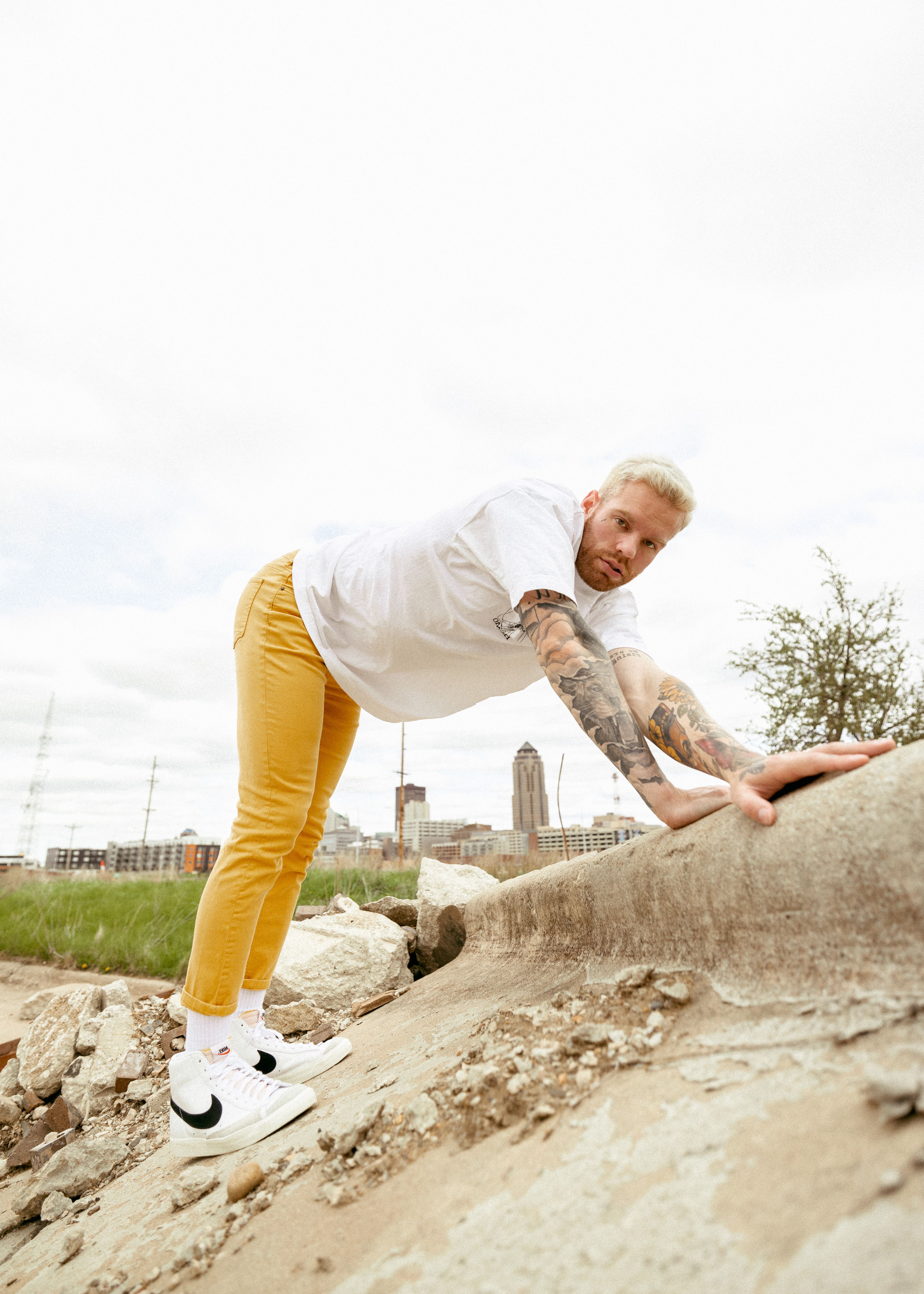 A tattooed man leaning on rocks with a city skyline in the background, outdoors.