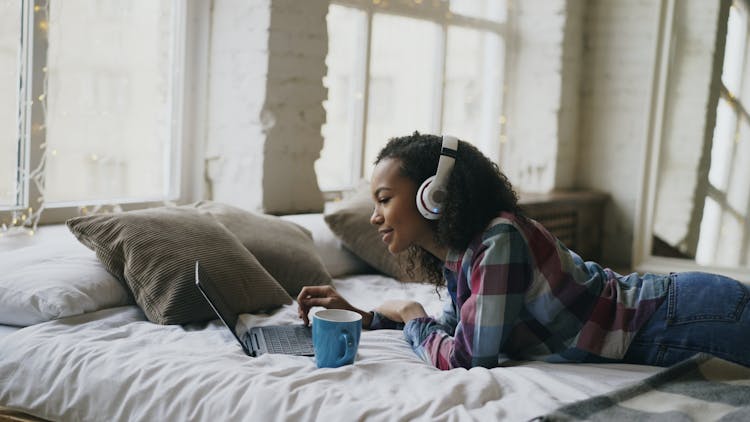 Smiling Girl In Headphones Relaxing On Bed With Laptop 