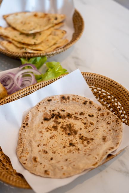 A stack of chapati, a charred naan, and a flaky paratha arranged on a dark wooden board with a small bowl of ghee alongside.