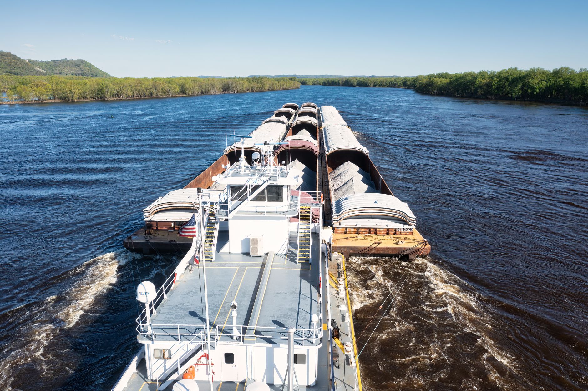 A large cargo barge navigating the Mississippi River in Wabasha, Minnesota, under clear skies.