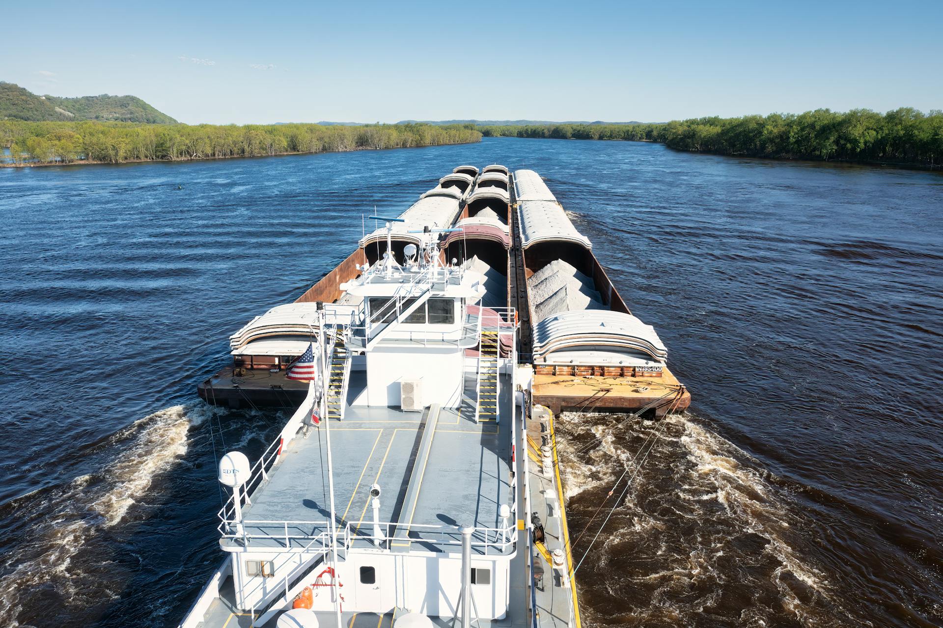 A large cargo barge navigating the Mississippi River in Wabasha, Minnesota, under clear skies.