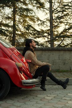 Young man with long hair leans against a classic red car surrounded by tall trees.