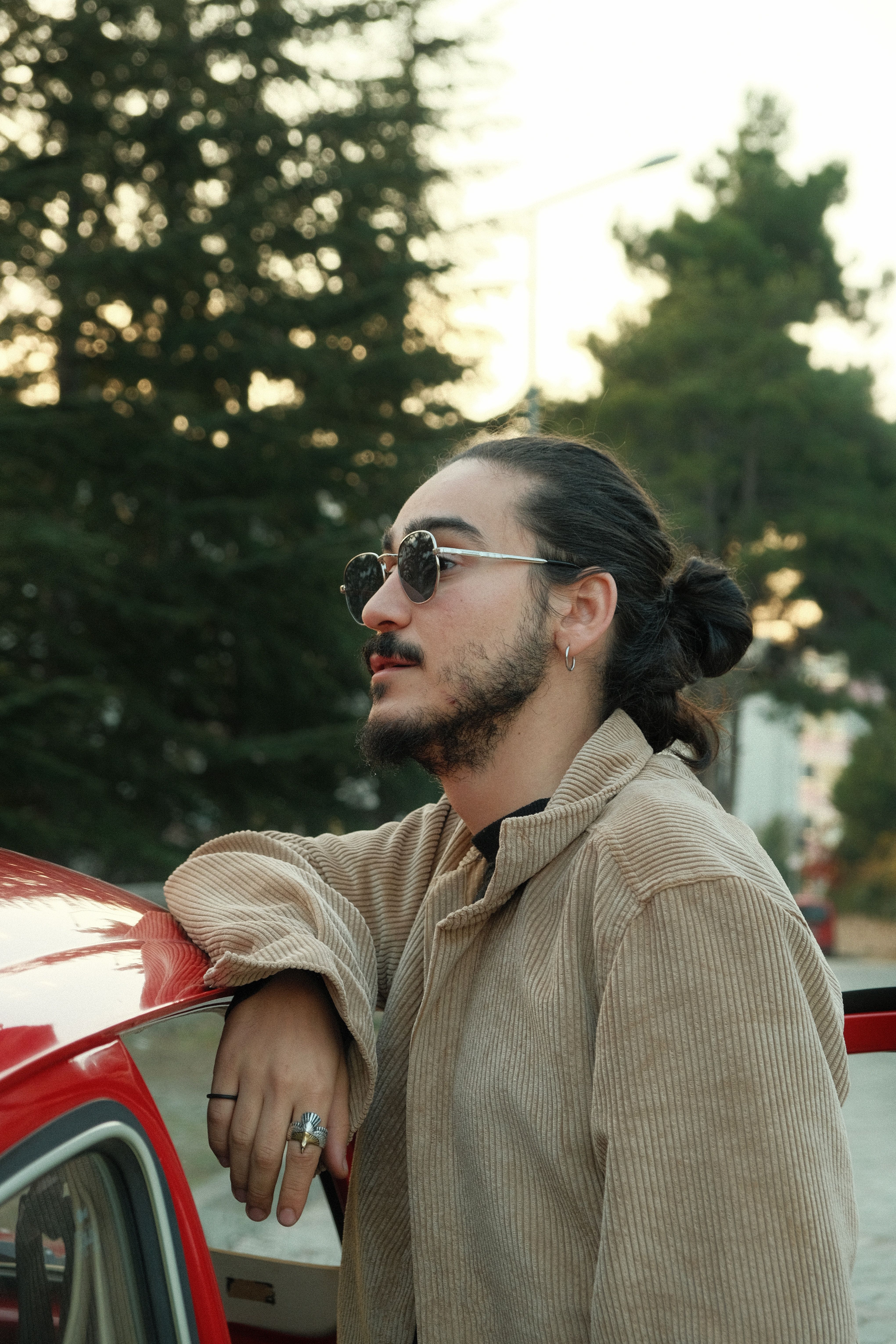 A stylish bearded man with sunglasses and man bun leans on a car. Relaxed and modern vibe.
