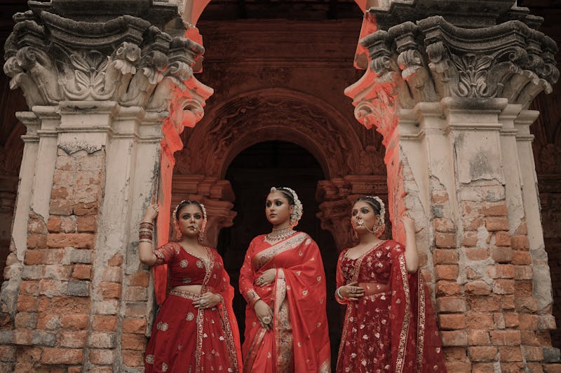 Three women in vibrant red sarees pose elegantly by ancient architecture.