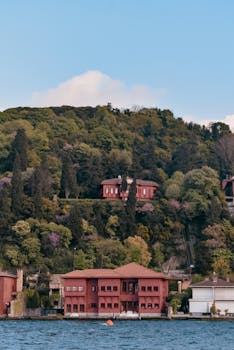 Picturesque red mansions nestled in lush forested hills beside a serene lakeshore under a blue sky.