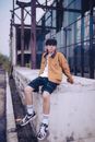 Man in Brown Jacket and Blue Denim Shorts Sitting on Concrete Wall