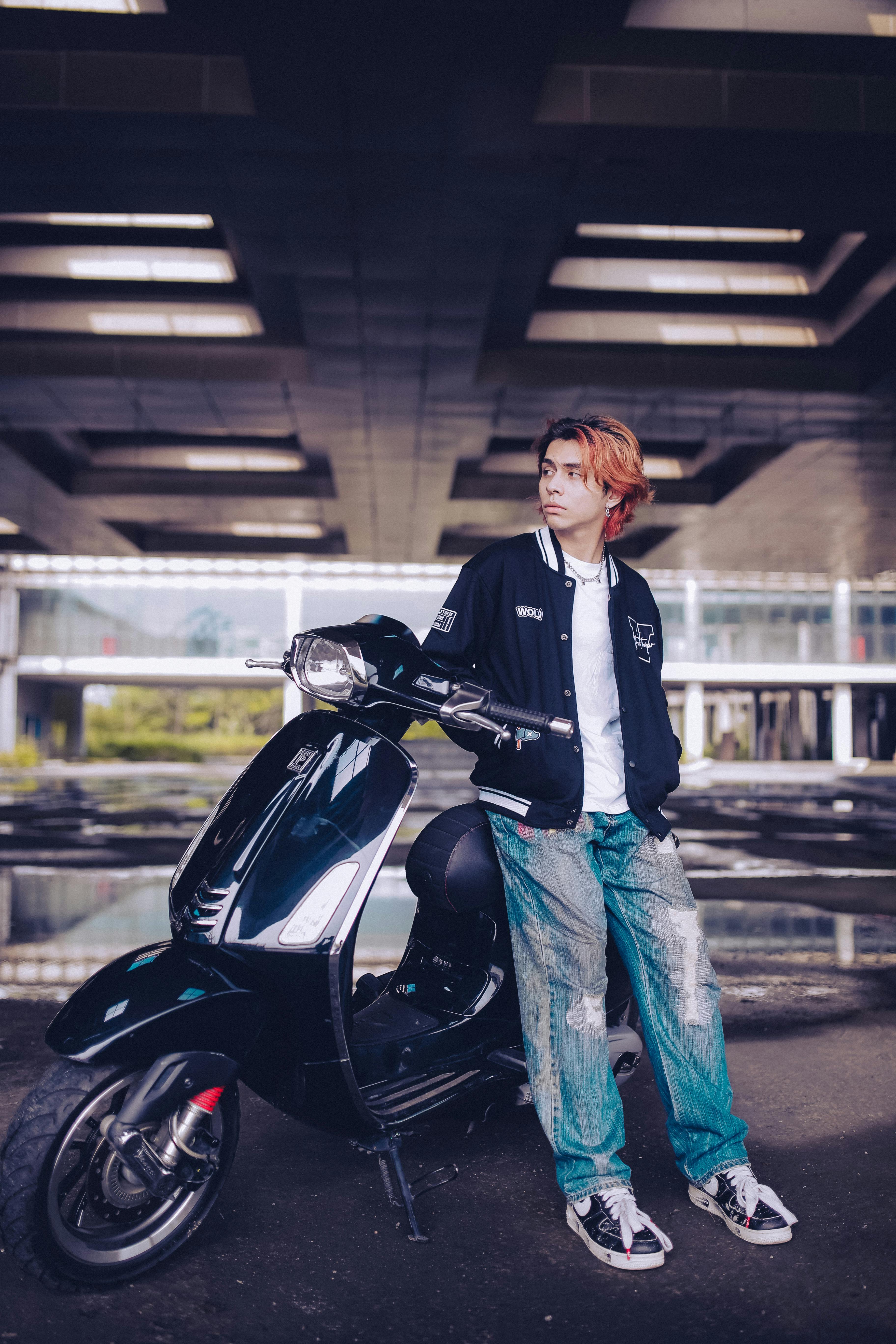 Stylish young adult stands confidently beside a sleek black scooter under a modern architectural canopy.