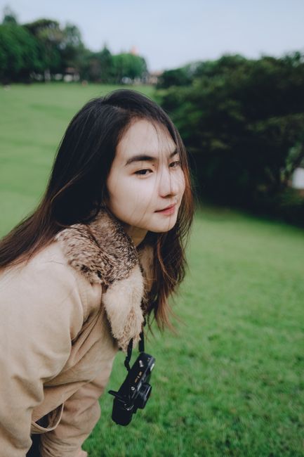Asian woman in fur coat with a DSLR camera enjoying a grassy park field.