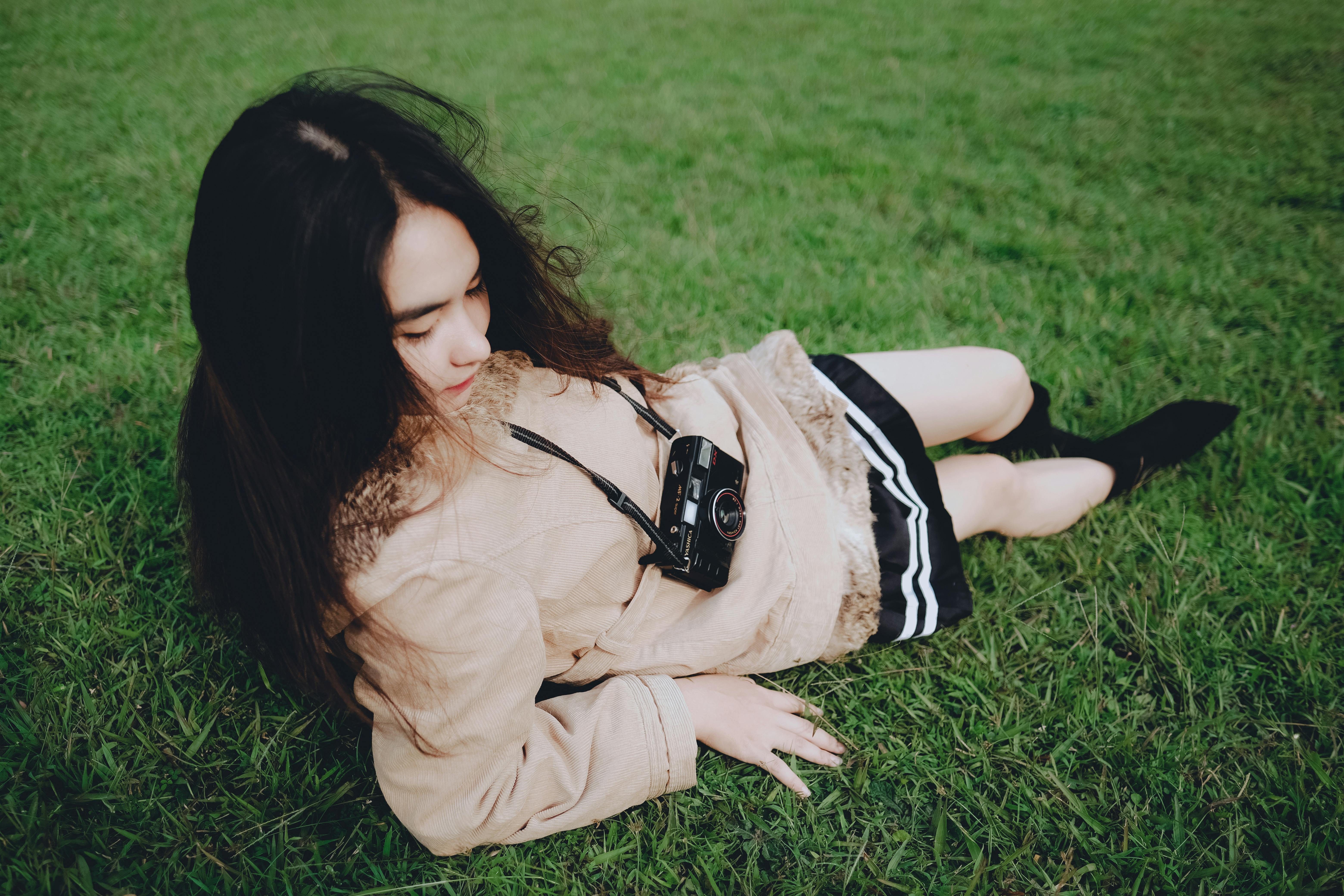 A woman lies on the grass outdoors, posing with a vintage camera, exuding a serene and fashionable vibe.