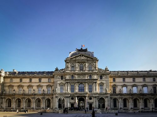 Free Front view of the iconic Louvre Museum in Paris under a clear blue sky with tourists outside. Stock Photo