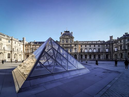 Free The Louvre Museum's glass pyramid in Paris, surrounded by historic architecture under a clear blue sky. Stock Photo