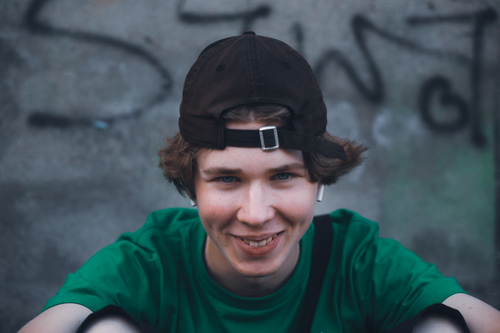 Free A joyful young man wearing a green shirt and brown cap, sitting outdoors against a graffiti wall. Stock Photo