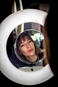 A woman gazes thoughtfully into a round mirror, framed by modern lighting.