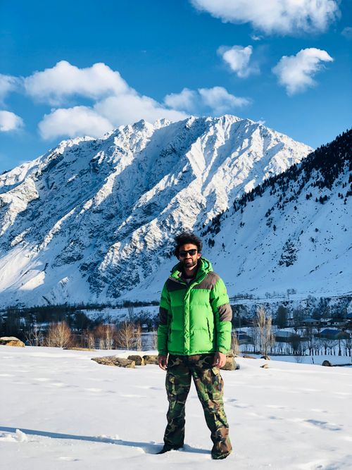 Free A man stands in snowy terrain, wearing a green jacket, with majestic snow-capped mountains as the backdrop. Stock Photo