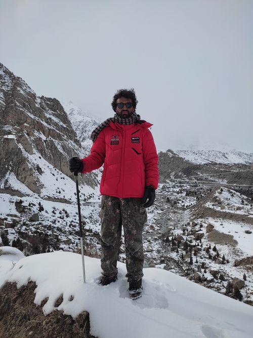 Free Man in red jacket stands on a snowy mountain holding a walking stick during winter adventure. Stock Photo
