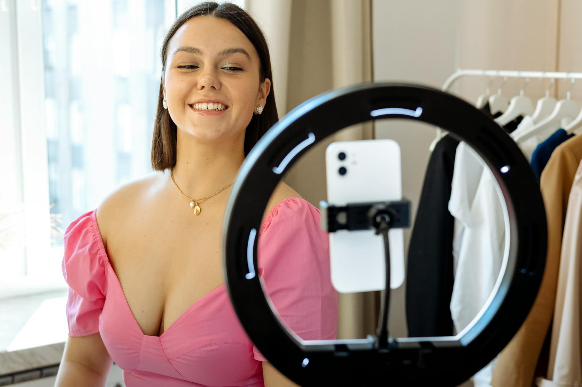 A fashionable young woman smiles while recording content using a smartphone and ring light at home.