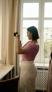 A young woman in a pink top taking photos inside a room with natural light.