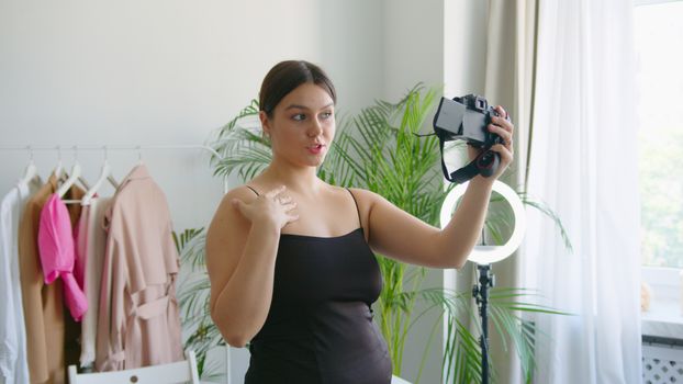 A young woman records a vlog indoors with a camera, surrounded by stylish clothing and plants.