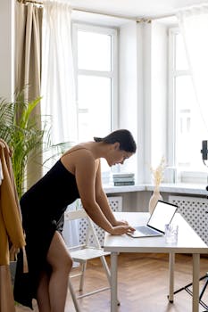 Woman in black dress using laptop in bright home office, emphasizing modern remote work style.
