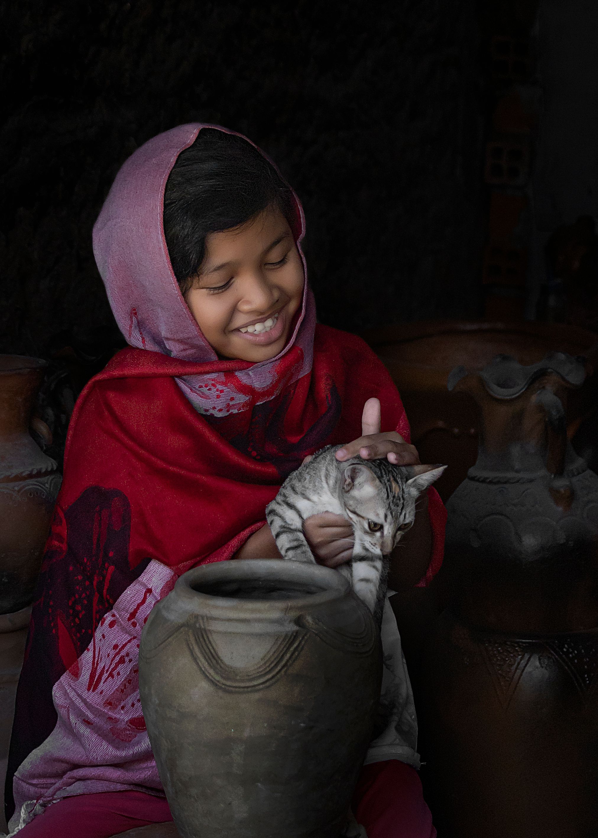 A cheerful girl wearing a red scarf holds a cat near a ceramic jar, creating a warm and intimate scene.