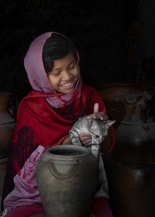 A Girl Sitting Near Gray Ceramic  Jar while  Looking at the Gray Cat she is Holding
