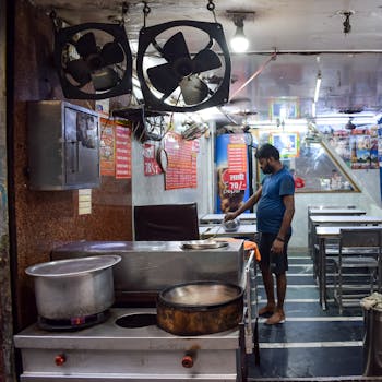 A man cooking in an indoor street food setting with industrial fans and tables.