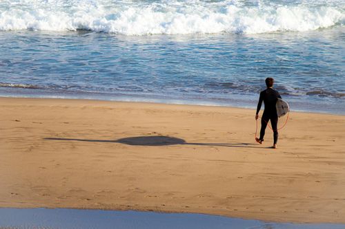 Free A surfer in a wetsuit walking towards ocean waves on a sunny day, carrying a surfboard. Stock Photo