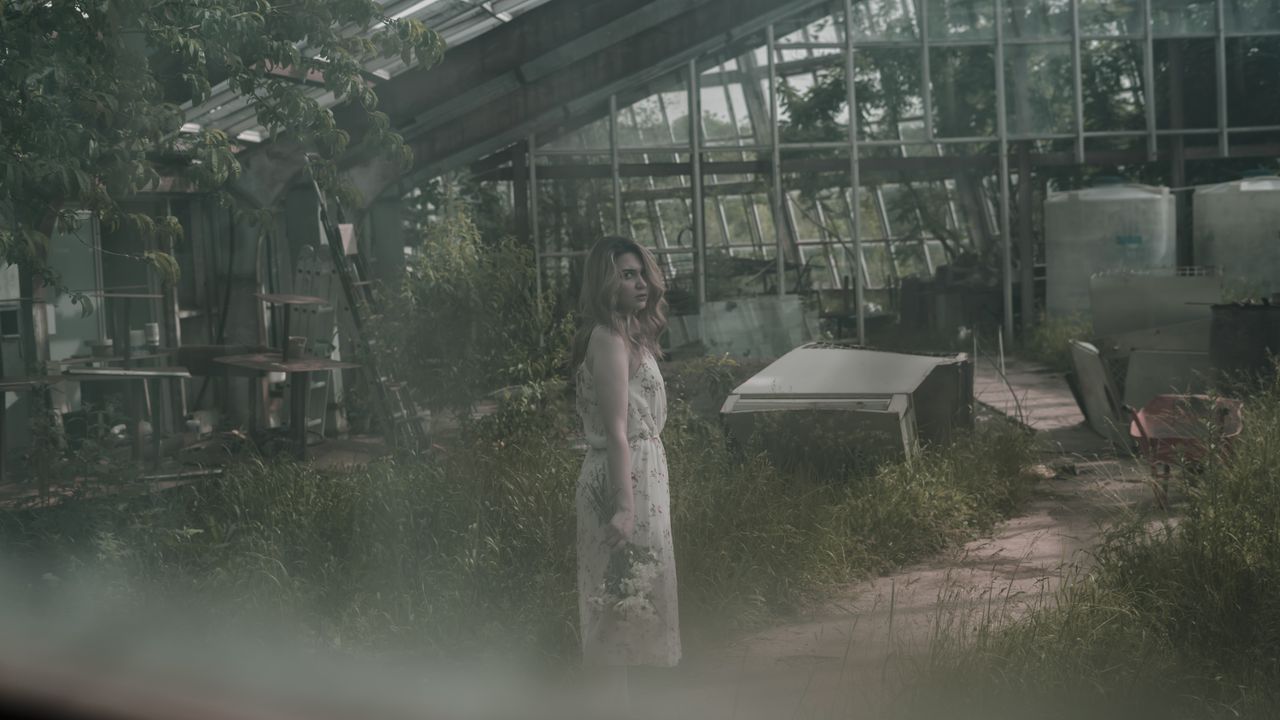 a woman in a dress stands in an overgrown abandoned greenhouse holding