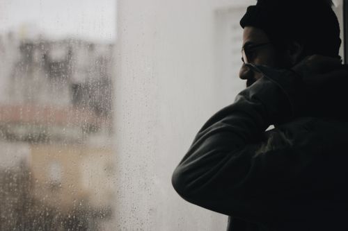 Free A man in a beanie gazes outside through a rainy window, creating a moody atmosphere. Stock Photo