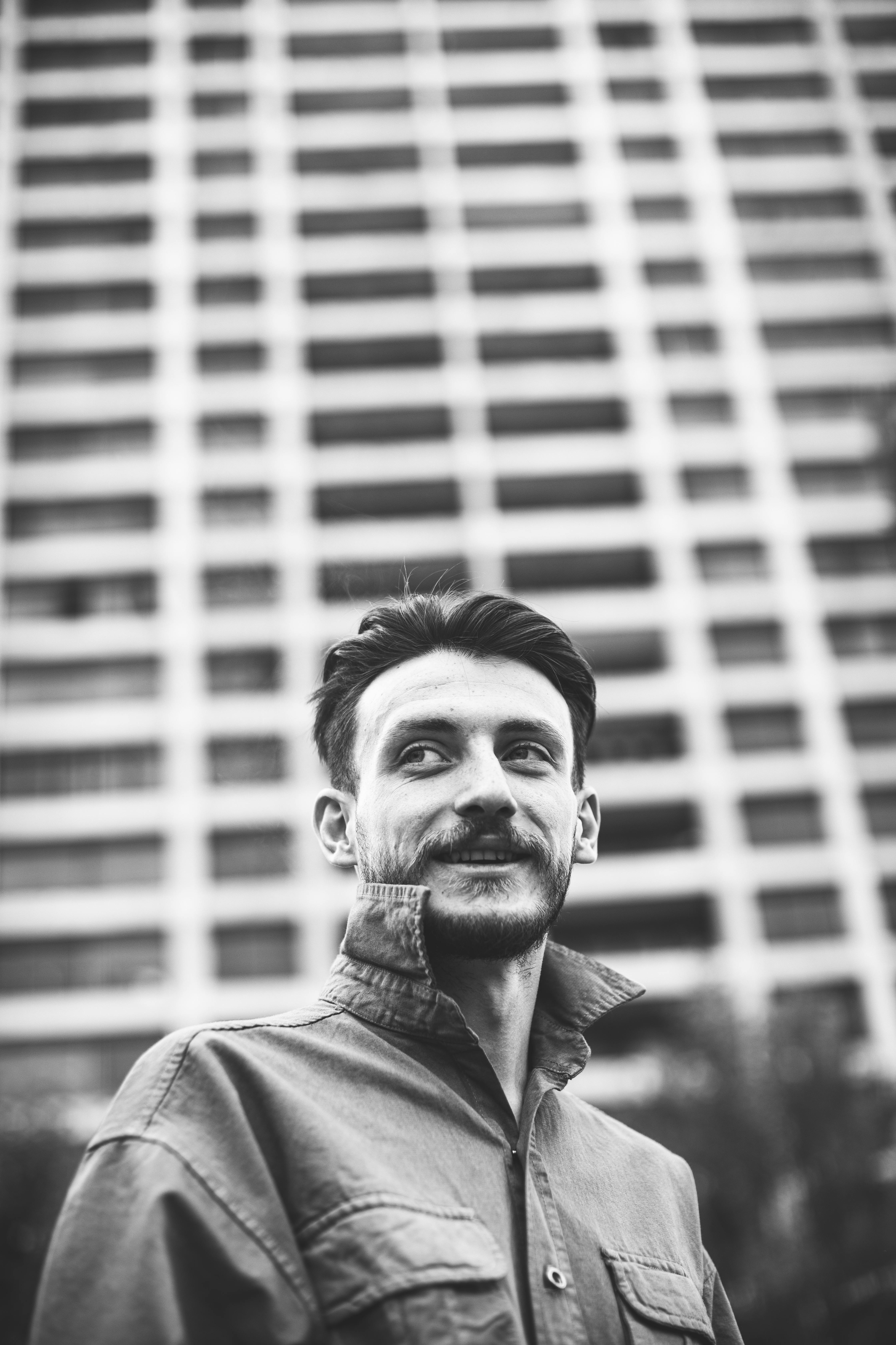Black and white portrait of a man with facial hair against a blurred building background.