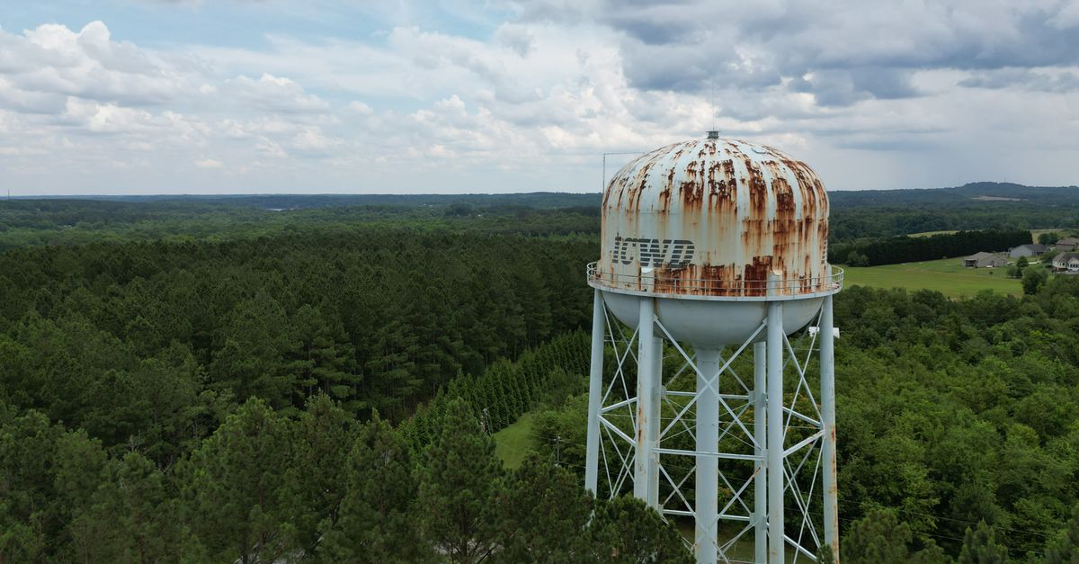 Drone shot of rusty water tower amidst lush green trees under cloudy sky in Campobello, SC.