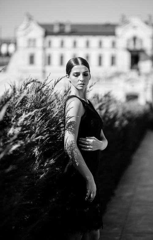 Free Stylish woman in black dress posing beside greenery with elegant architecture in background. Stock Photo