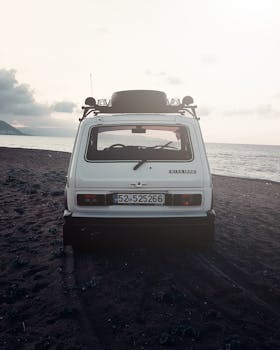 A classic white car parked on a sandy beach with the serene sea and sunset in the background.
