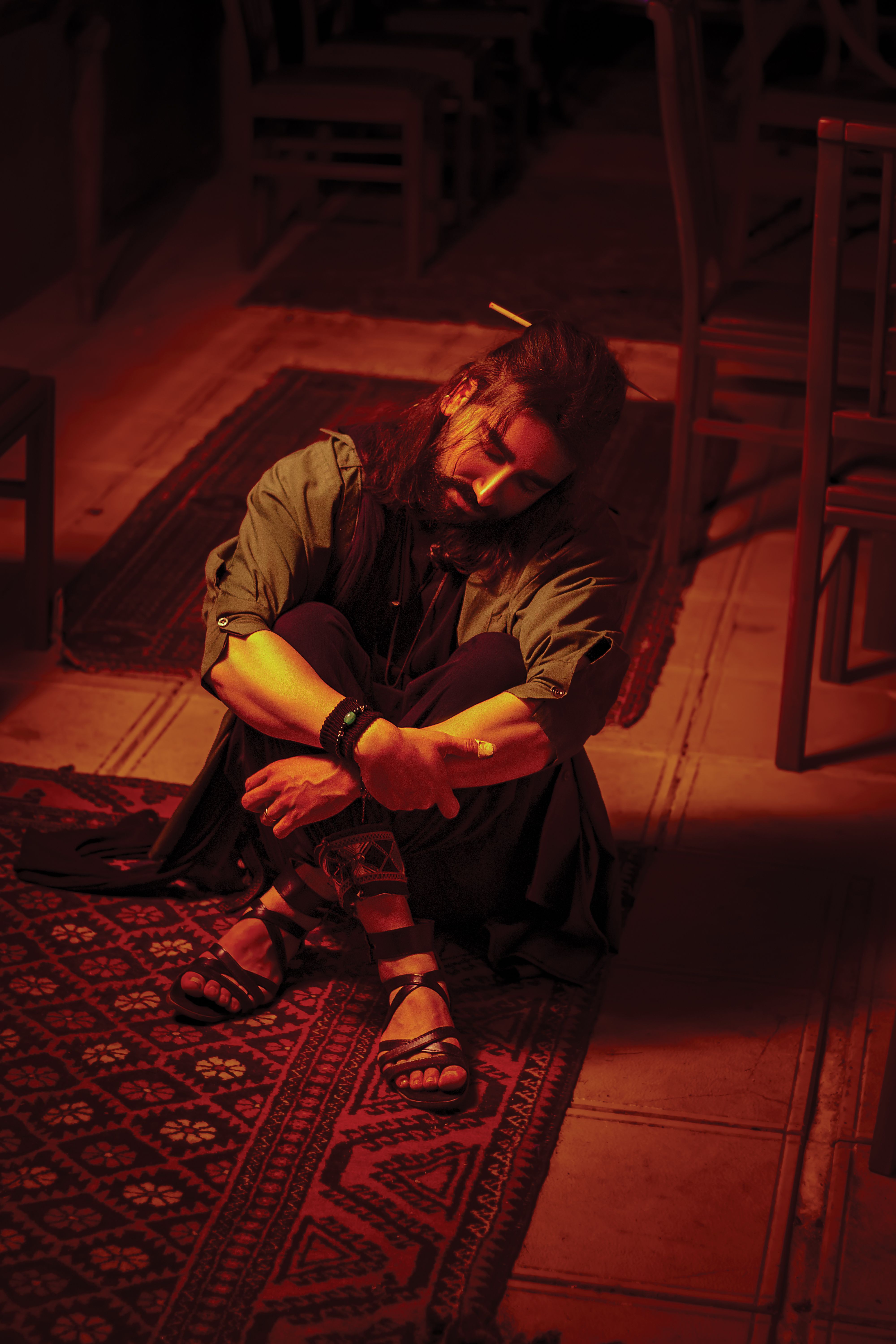 A man sits contemplatively on a patterned rug in warm, dim lighting, creating a moody atmosphere.