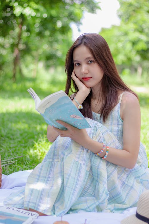 Free Shallow Focus of a Pretty Woman in Checkered Dress Sitting on Picnic Blanket while Holding a Book Stock Photo