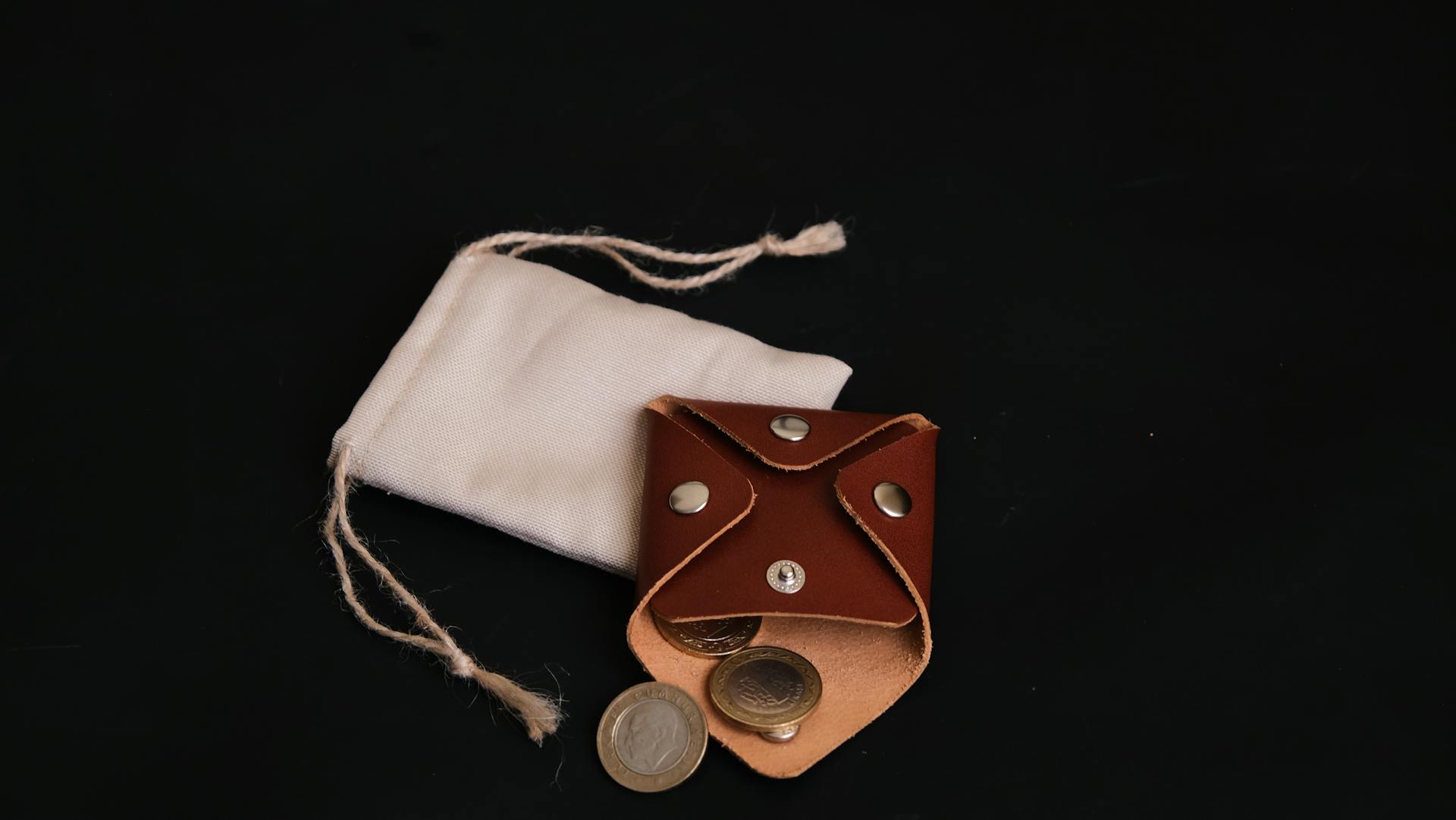 Brown leather coin purse with coins and white cloth bag on black background.