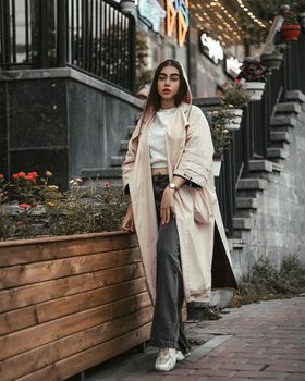 Fashionable woman in long coat posing near flowers and stairs in city.